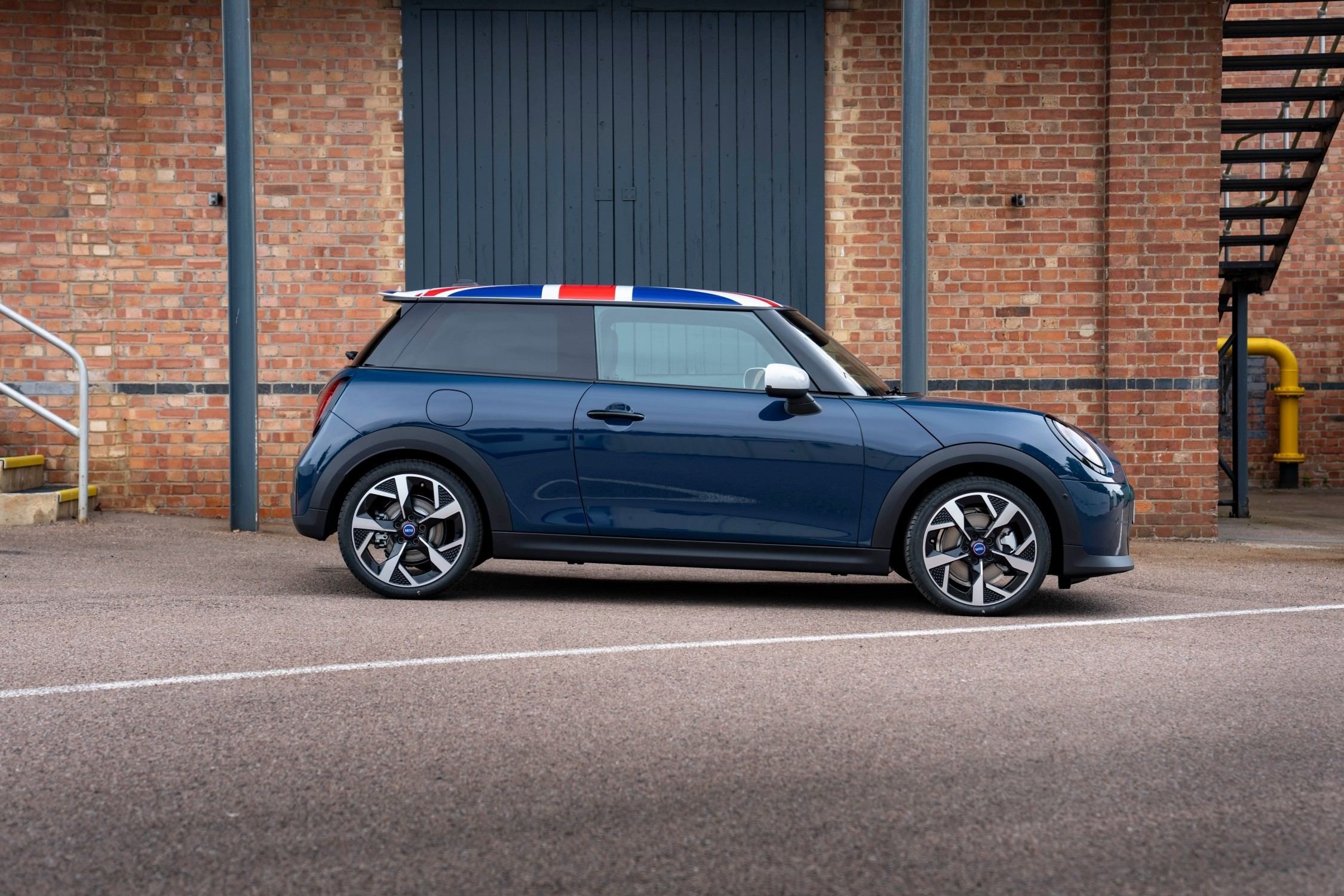 MINI Cooper Oxford Edition azul visto de perfil, com tejadilho Union Jack e capas dos espelhos retrovisores em branco, estacionado em frente a um edifício de tijolo